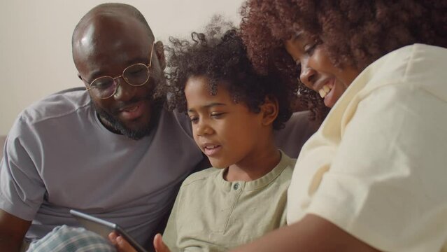 Cheerful African American parents and little son sitting together on sofa at home and discussing something on digital tablet