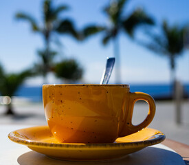 yellow cup of coffee or tea with spoon on the table, in the sunshine, with blue sky and palm trees in the background