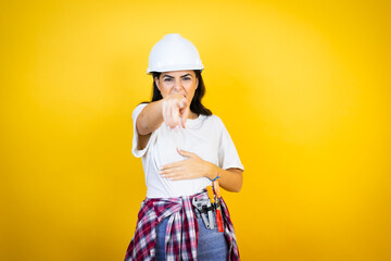 Young caucasian woman wearing hardhat and builder clothes over isolated yellow background laughing at you, pointing finger to the camera with hand over body, shame expression
