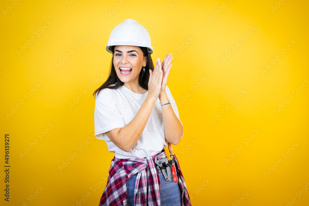 Wall mural Young caucasian woman wearing hardhat and builder clothes over isolated yellow background clapping and applauding happy and joyful, smiling proud hands together