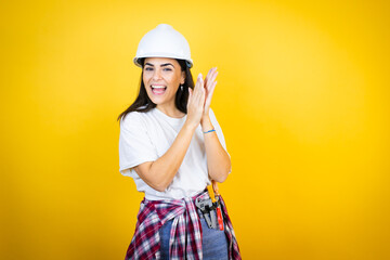 Young caucasian woman wearing hardhat and builder clothes over isolated yellow background clapping...