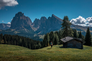 Fototapeta na wymiar Girl hiking along meadows with wooden cabin at Alpe di Siusi during summer with view to mountains of Plattkofel and Langkofel in the Dolomite Alps in South Tyrol, Italy.