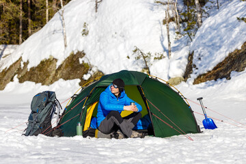 Happy man drinking tea in a green tent in the forest. Nearby are a backpack and a snow shovel....