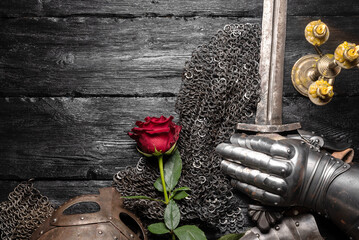Knight sword, red rose flower and iron helm on the black woorden flat lay table background.