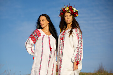 Two Pretty young ladies wear traditional Ukrainian clothes and flower wreath walk in wheat field, beautiful ethnic girl in handmade decorated floral crown admire nature. Sunset.