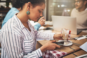 Always busy as usual. Shot of a focused young woman texting on her cellphone while being seated at a coffeeshop drinking a hot beverage.
