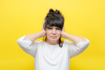 elderly woman with an expression of pain on her face covers her ears with her hands, on a yellow background, the concept of pain, age, unhappiness, unwillingness to hear