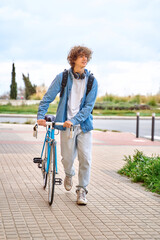 Young curly-haired student with headphones walking to college holding bicycle with hands.