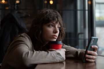 A girl with curly hair uses a smartphone while sitting in a cafe by the window.