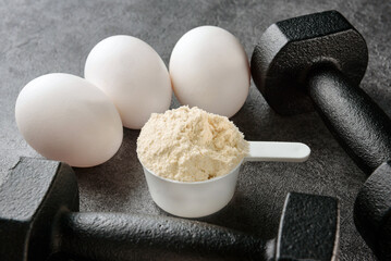 A scoop of egg white, eggs and a pair of black dumbbells on a dark background close-up.