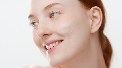 Extreme close-up beauty portrait of young ginger Caucasian woman with cream smear on her cheek smiles wide looking aside on white background | Face care commercial concept