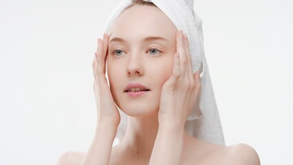 Young ginger Caucasian woman with a hair drying towel on her head touches her face with both hands massaging her temples looking aside on white background | Beauty portrait