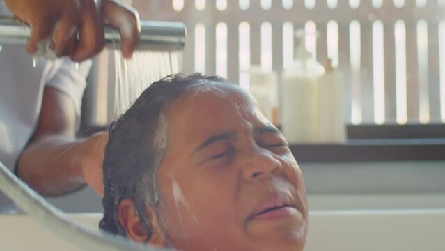 Tilt up shot of little African American boy sitting with eyes closed in bath while father rinsing shampoo out of his hair with shower