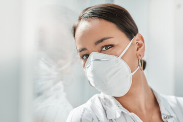 2020 The year of the face mask. Shot of a masked young businesswoman leaning against a window in a modern office.