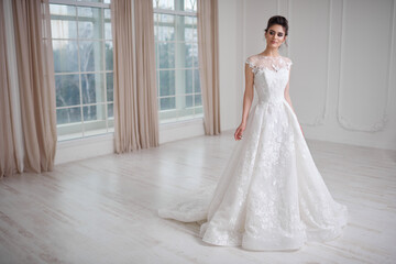Full length image of a young attractive bride in beautiful dress sitting posing indoors in white studio with windows.