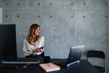 The smiling caucasian woman is smiling and holding a smartphone in her hands. She's sitting in the office chair and looking through the window.