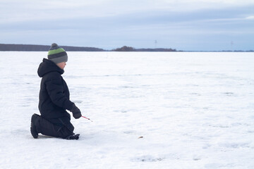 Fototapeta na wymiar A boy catches fish in winter with a fishing rod on the ice of the river on a winter fishing trip