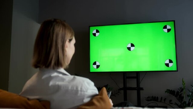 A girl is sitting at home in the living room and watching TV with a green screen Chroma Key while relaxing on the sofa. Changes TV channels with the remote control.