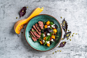 Baked pumpkin salad, spinach, tomato and beef in a white plate on a stone background, top view