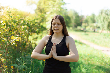 Naklejka na ściany i meble A brunette in a sporty black T-shirt folded her arms on her chest in a namaste gesture. Yoga classes in the fresh air. Group yoga practices during quarantine in the park