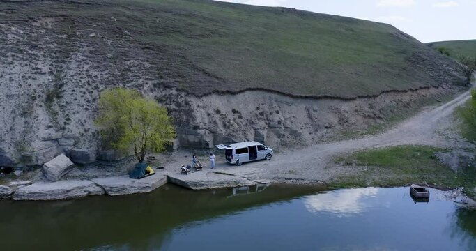 the drone flies up in the gorge of the lake river to the campsite where the guy and the girl are resting near the car and the tent. the guy controls the drone, the girl waves her hand