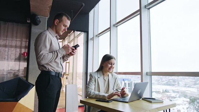 Pair of colleagues in office. Woman sits at desk in front of laptop addressing man. Male workmate looks at the phone in his hands.