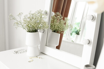 Vase with gypsophila flowers and jewelry on dressing table near light wall