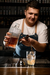 Young bartender pours liquid from jigger into crystal mixing cup