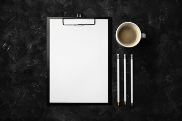 Photo of clipboard with blank paper, coffee cup and pencils on black plaster background. Office...