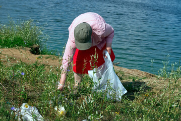 An elderly woman cleans the natural area of garbage, collects waste on the river bank in a large bag.