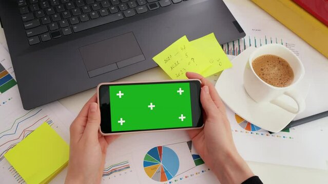 Business woman looking at green screen Mock-up Chroma Key of smartphone sitting at desk in business office, POV. Top view female using phone, copy space, worksheet finance graph account chart market p