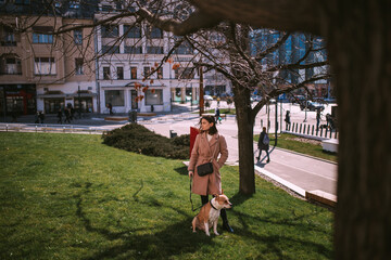 Caucasian young girl poses with her brown white Stafford in green area in city, under the tree. She looks on her right side and dog looks on its left. Stafford is on a leash. She wears coat and purse.