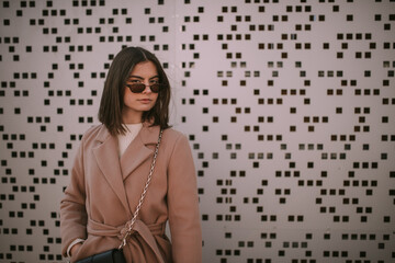 A beautiful, young, fashionable caucasian girl in a beige coat poses in front of the checkered background. She wears sunglasses, a white blouse, black purse with gold chine. She looks right at camera.