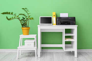Workplace with modern laptop, printer and stepladder stool with houseplant near green wall in room interior