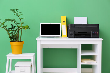Workplace with modern laptop, printer and stepladder stool with houseplant near green wall