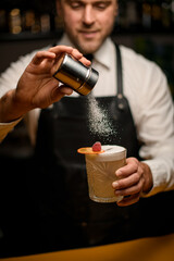 bartender gently holds glass full of foamy cocktail and sprinkles on it with sugar powder