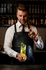 barman gently holds and sprinkles glass of cold cocktail decorated with mint twig and cucumber stripes