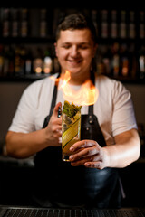 Young brunette bartender sets fire to the transparent alcohol drink with cucumber, mint and ice