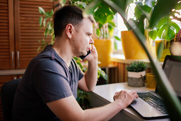 A man working with laptop remotely from home. A distant work place with many home plants. Green...