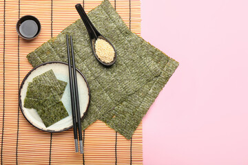 Plate of tasty seaweed sheets, spoon with sesame, bamboo mat, soy sauce and chopsticks on pink...