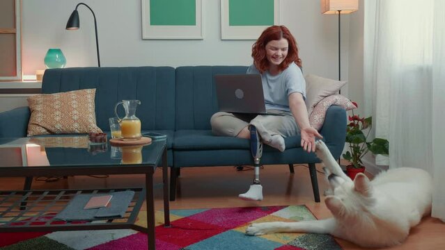 Attractive happy disabled woman with bionic prosthetic leg using laptop at home working online, petting big white dog.
