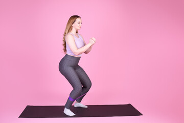 A beautiful body-positive girl on a pink background in a full-length sports suit goes in for sports, elastic bands for training, leg exercises, fitness