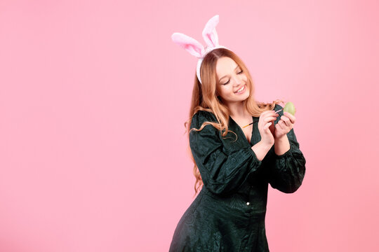 A beautiful girl in bunny ears paints Easter eggs on a pink background. Woman smiling and laughing, model with beautiful hair in green dress.