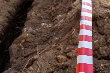 Red and white barrier tape protect a danger place. The warning tape indicates the danger of being on the territory of the construction site. Dug trenches for laying new pipes.