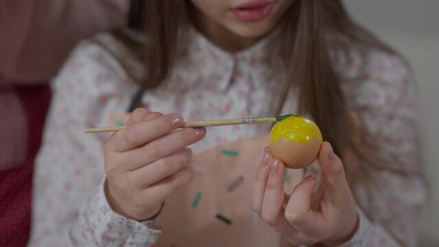 Close-up boiled egg in hands of unrecognizable teenage girl painting with brush on food. Concentrated Caucasian teenager helping parent preparing traditional dinner on holiday at home indoors