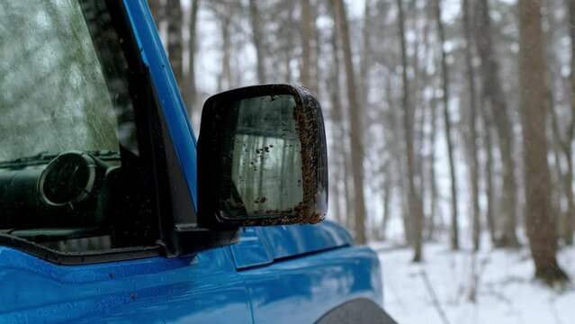 Dirty blue side view mirror of SUV in snowy forest. It's snowing, ride on loose earth, mud flew from under wheels and stained car body.