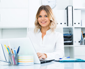 Smiling girl in white shirt sitting at office desk in well-lit office. High quality photo