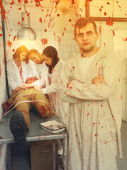 Portrait of young man with friends standing in quest room with bloody traces on walls and zombie mannequin on surgical table