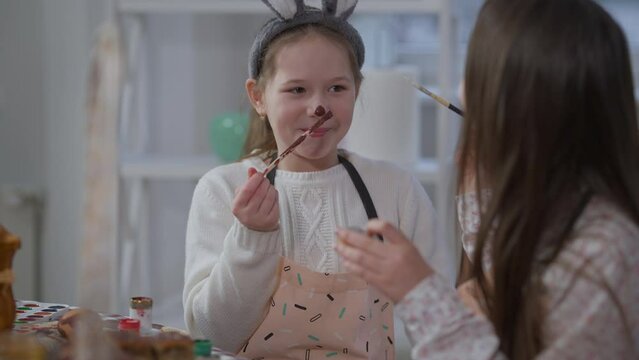 Charming joyful girl coloring nose with food paint sitting at table with sister. Portrait of cheerful cute Caucasian child having fun preparing Easter dinner with sibling at home