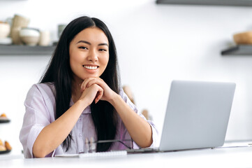 Lovely positive Asian brunette girl in a stylish shirt, designer, freelancer or student, sits at home in the kitchen with a laptop, working or study remotely, looking at the camera, smiling friendly - obrazy, fototapety, plakaty